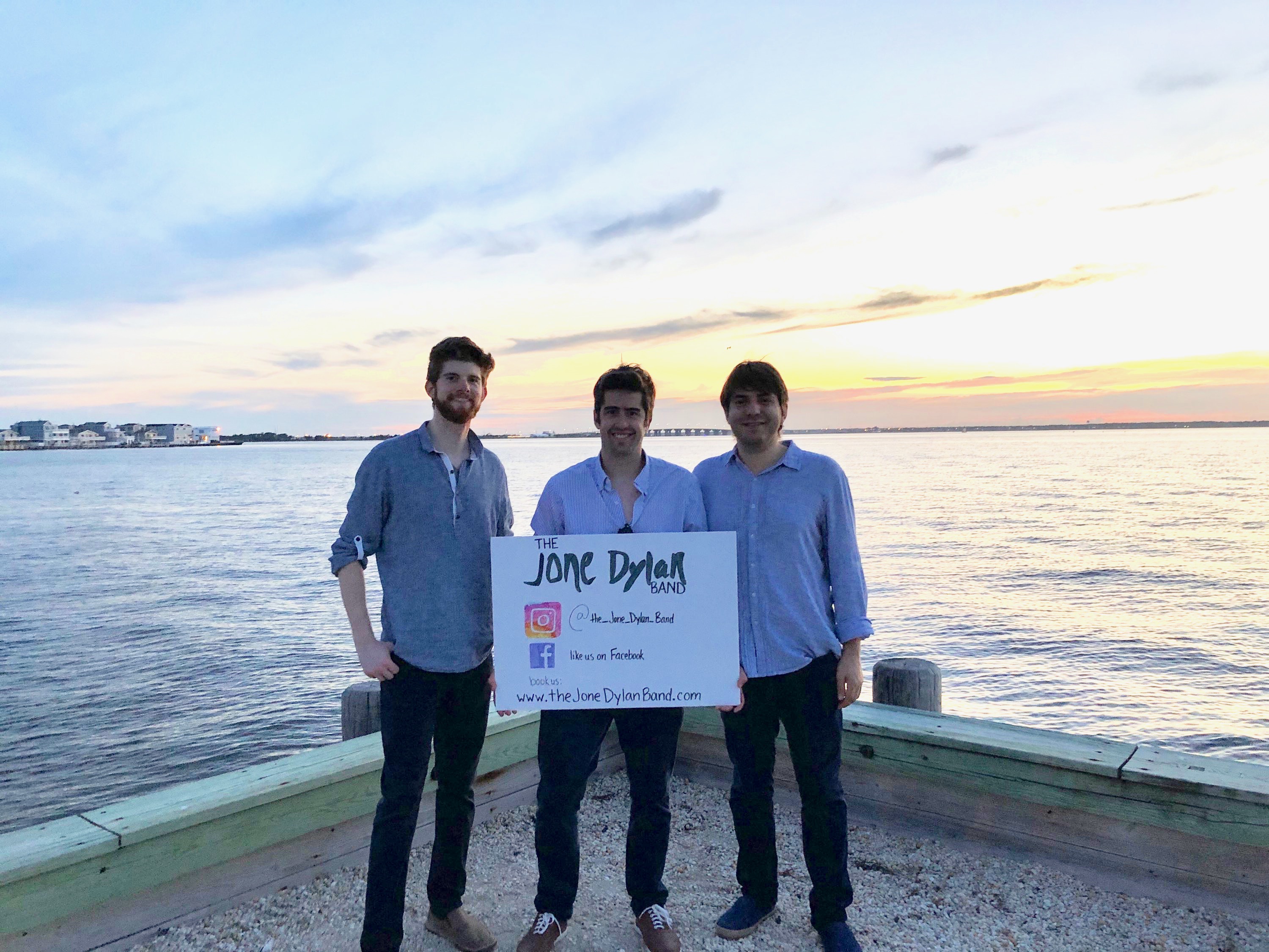 Jone Dylan, DiGiac, and Nikitas at the Surf City Yacht Club with a poster containing the band logo.